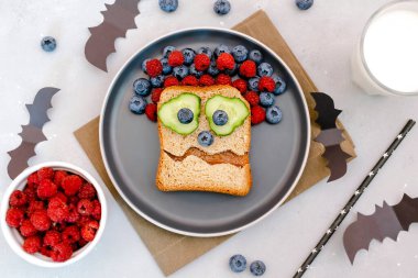 Funny scary monster face smile on halloween sandwich toast bread with peanut butter, blueberry, raspberry on plate,bats on gray background close up. Kids child sweet dessert breakfast lunch food.