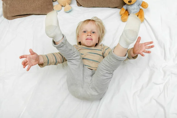 Cute little blond caucasian child kid toddler boy playing,having fun, lying on bed,lifts up the legs at home at sunny morning wakes up in his room. happy childhood moments.
