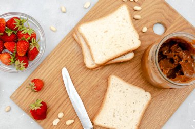 Fıstık ezmeli tost ekmeği, çilek, tahta kesme tahtasında fındık. Çocukların tatlıları, sağlıklı kahvaltı, öğle yemeği, gri arka planda yemek sanatı, üst manzara, düz yatak..