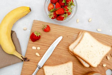 Fıstık ezmeli tost ekmeği, muz, çilek, ahşap kesim tahtasında fındık. Çocukların tatlıları, sağlıklı kahvaltı, öğle yemeği, gri arka planda yemek sanatı, üst manzara, düz yatak..