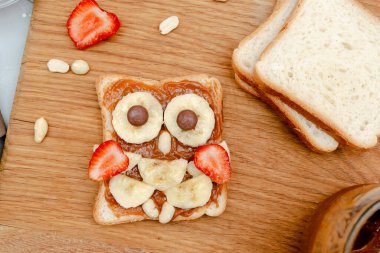 Komik baykuş suratlı tost ekmeği, fıstık ezmesi, muz, ahşap tahtada çilek. Tatlı çocuklar bebeğin tatlı tatlısını, sağlıklı kahvaltısını, öğle yemeğini, gri arka planda yemek sanatını, üst manzarayı....