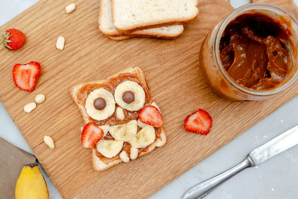 Funny owl face sandwich toast bread with peanut butter, banana, strawberry on wooden board. Cute kids childrens baby\'s sweet dessert, healthy breakfast,lunch, food art on gray background,top view.