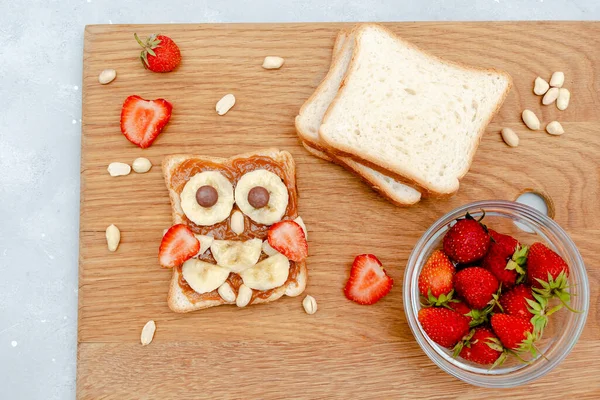 Funny owl face sandwich toast bread with peanut butter, banana, strawberry on wooden board. Cute kids childrens baby\'s sweet dessert, healthy breakfast,lunch, food art on gray background,top view.