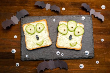 Cadılar bayramında komik canavar suratları yumuşak yeşil avokado peynirli ricotta tereyağlı tost ekmeği servis tahtasında salatalıklar, tahta zeminde yarasalar. Çocuklar için öğle yemeği..