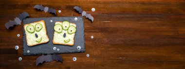 Cadılar bayramında komik canavar suratları yumuşak yeşil avokado peynirli ricotta tereyağlı tost ekmeği servis tahtasında salatalıklar, tahta zeminde yarasalar. Çocuklar için öğle yemeği..