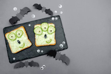 Funny monster faces on halloween sandwich toast bread with soft green avocado cheese ricotta butter,cucumbers on cutting serving board,bats on gray background.Kids child breakfast lunch food.
