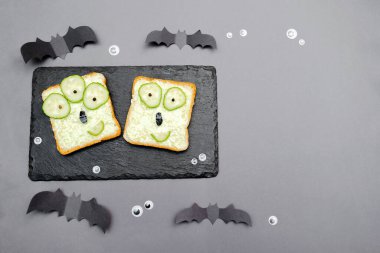 Funny monster faces on halloween sandwich toast bread with soft green avocado cheese ricotta butter,cucumbers on cutting serving board,bats on gray background.Kids child breakfast lunch food.
