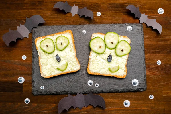 Cadılar bayramında komik canavar suratları yumuşak yeşil avokado peynirli ricotta tereyağlı tost ekmeği servis tahtasında salatalıklar, tahta zeminde yarasalar. Çocuklar için öğle yemeği..