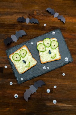 Cadılar bayramında komik canavar suratları yumuşak yeşil avokado peynirli ricotta tereyağlı tost ekmeği servis tahtasında salatalıklar, tahta zeminde yarasalar. Çocuklar için öğle yemeği..