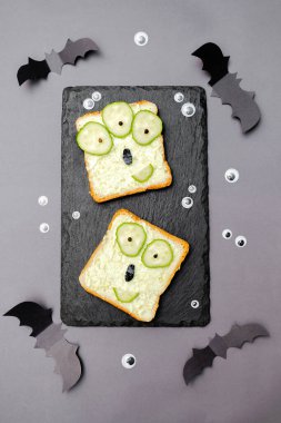 Funny monster faces on halloween sandwich toast bread with soft green avocado cheese ricotta butter,cucumbers on cutting serving board,bats on gray background.Kids child breakfast lunch food.