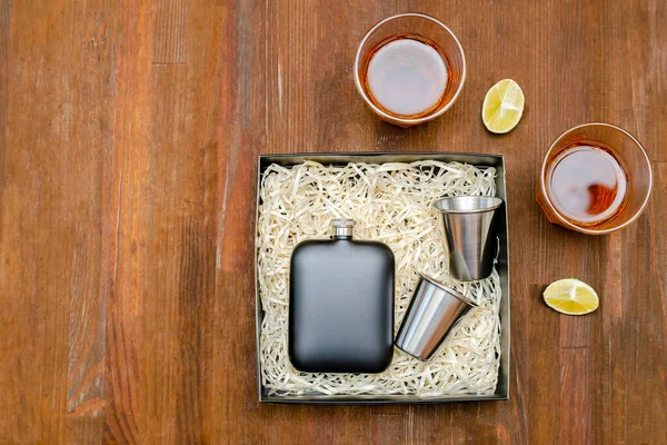 ステンレス製の黒金属製のポケットフラスコ ギフトボックスハイキングマンは メガネ付きアルコールのために設定します ウイスキーのグラス木製のテーブルの背景にコニャックドリンクトップビューコピースペースモックアップフラットレイ — ストック写真