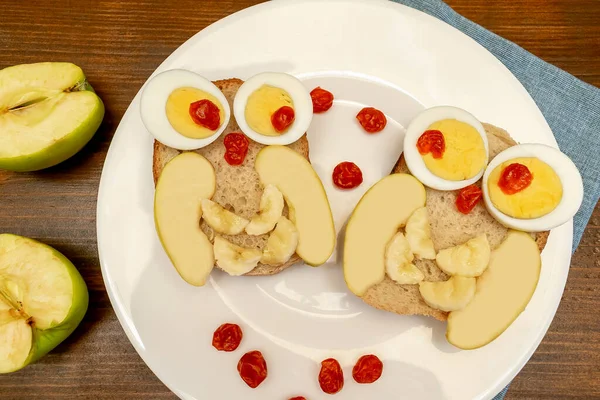 Funny owl face sandwich toast bread with chicken eggs, apple, banana, dried berry fruits on plate. Cute kids childrens baby's sweet dessert breakfast,lunch, food art on wooden background,top view.
