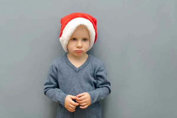 Unhappy gloomy sad cute little child kid baby boy in christmas red santa claus hat cap in front of gray wall in room,celebrating New Year at home. Merry Christmas and Happy Holidays.mock up