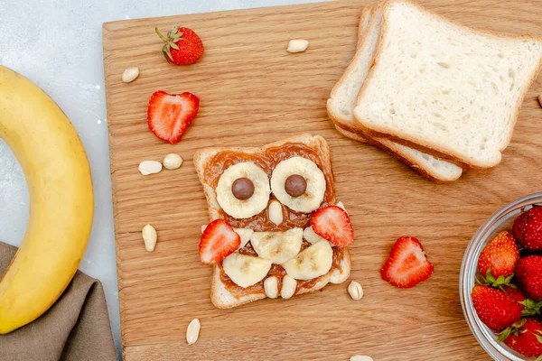 Funny owl face sandwich toast bread with peanut butter, banana, strawberry on wooden board. Cute kids childrens baby\'s sweet dessert, healthy breakfast,lunch, food art on gray background,top view.