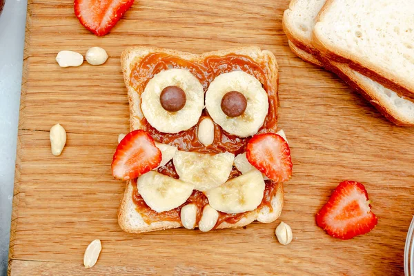 Funny owl face sandwich toast bread with peanut butter, banana, strawberry on wooden board. Cute kids childrens baby\'s sweet dessert, healthy breakfast,lunch, food art on gray background,top view.