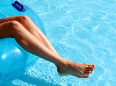 Havuz parkında şişme yüzme çemberinde oturan narin kadın ayaklı kız resmi var. Havuzda mavi şeffaf su.