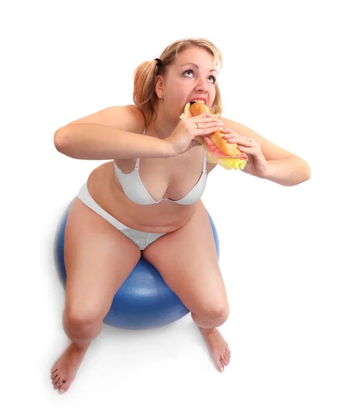 Mulher com excesso de peso sentada em uma bola de fitness comendo sanduíche grande . — Fotografia de Stock
