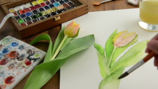 Painting with paintbrush orange tulip bud in watercolors and cleaning the brush in water glass.