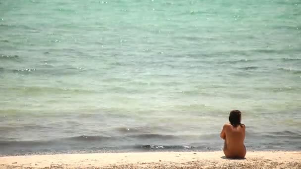 Egy nő ül a strandon, meztelen, tengerre néző — Stock videók