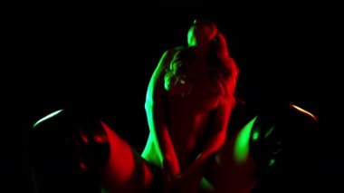 Güzel bir kadın çıplak poz veriyor. Neredeyse gölgede, üzerinde yumuşak mavi ve kırmızı ışıklar var.