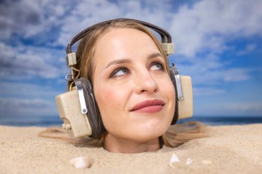 Kafasına kadar plaja gömülü güzel bir kadın kulaklıkla müzik dinliyor.