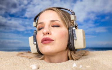 Kafasına kadar plaja gömülü güzel bir kadın kulaklıkla müzik dinliyor.