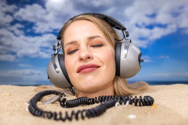 Kafasına kadar plaja gömülü güzel bir kadın kulaklıkla müzik dinliyor.
