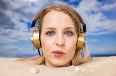 Kafasına kadar plaja gömülü güzel bir kadın kulaklıkla müzik dinliyor.