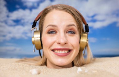 Kafasına kadar plaja gömülü güzel bir kadın kulaklıkla müzik dinliyor.