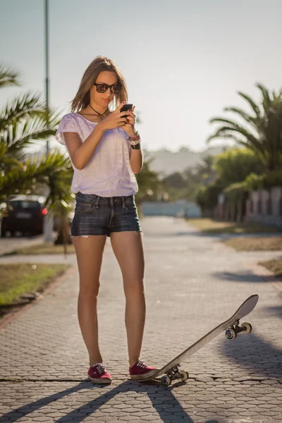 Молодая девушка со скейтбордом с помощью смартфона на открытом воздухе — стоковое фото