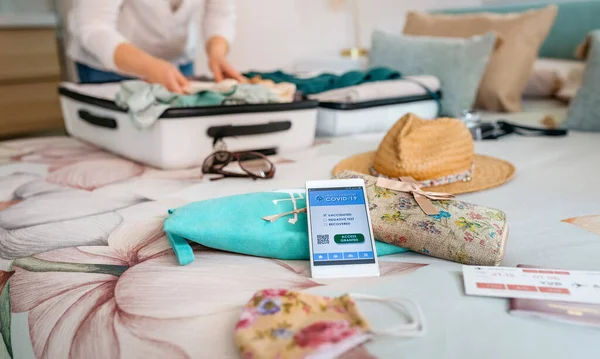 Teléfono celular con pasaporte covid con mujer irreconocible preparando la maleta para las vacaciones — Foto de Stock