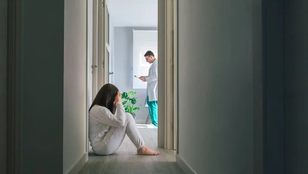 Paziente seduto sul pavimento nel corridoio di un centro di salute mentale con un medico sullo sfondo — Foto Stock