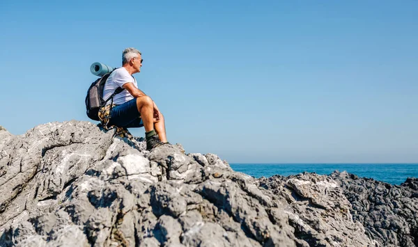 Homme âgé randonnée regardant le paysage marin assis sur des rochers — Photo