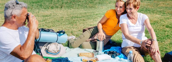 Padre anziano che scatta una foto alla sua famiglia seduta su una coperta mentre fa un picnic — Foto Stock