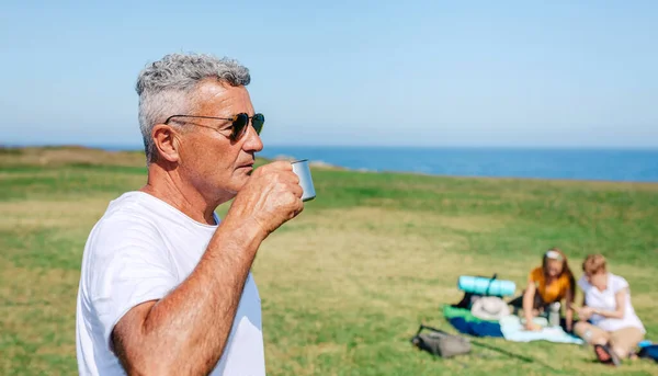 Senior man with sunglasses drinking coffee outdoors with his family in background — Stock Photo, Image