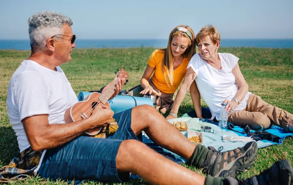 Senior man playing ukulele for his family sitting on a blanket during an excursion — Stock Photo, Image