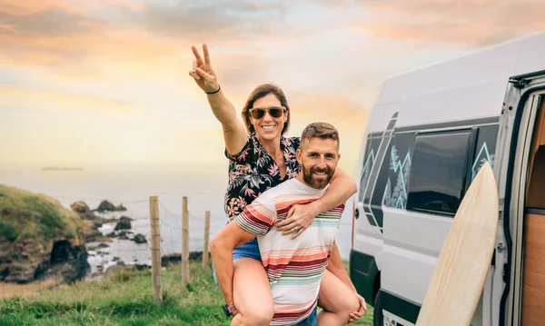 Paar schaut Kamera und hat Spaß Huckepack neben ihrem Wohnmobil — Stockfoto