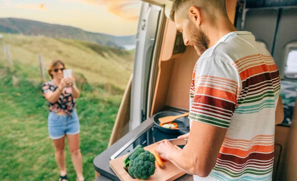 Jovem cozinhar legumes em uma van campista, enquanto sua esposa tira uma foto dele com seu telefone celular — Fotografia de Stock