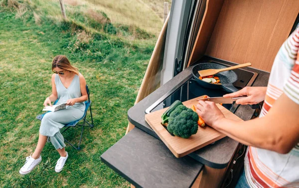 Uomo cucina verdure in un camper mentre la donna legge all'aperto — Foto Stock
