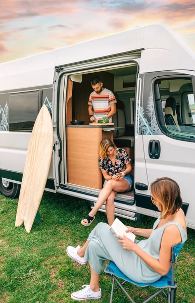 Amici che viaggiano in camper. Uomo cucina, donna lettura e donna alla ricerca mobile — Foto Stock