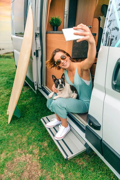 Young woman taking photo sitting with her boston terrier dog at the door of camper van