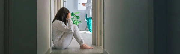 身穿睡衣的女病人坐在精神健康中心走廊的地板上 背后坐着一位医生 — 图库照片
