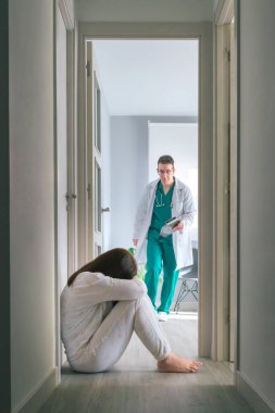 Worried physician running to help mental disorder female patient sitting on floor in mental health center hallway