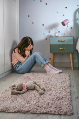 Unrecognizable sad girl with anxiety sitting on the floor of her bedroom with stuffed animal toy lying. Selective focus on stuffed animal in foreground