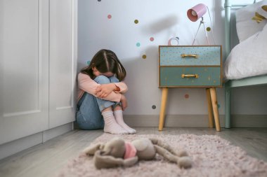 Unrecognizable sad little girl sitting on the floor of her bedroom with stuffed toy lying. Selective focus on girl in background