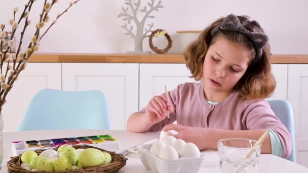 Ένα κοριτσάκι βάφει αυγά για το Πάσχα. Παιδί σχολιαρόπαιδο ευρωπαϊκής εμφάνισης και πασχαλινά αυγά, διακόσμηση στο τραπέζι. Ελαφρά εσωτερικά και φυσικά πλάνα. — Αρχείο Βίντεο