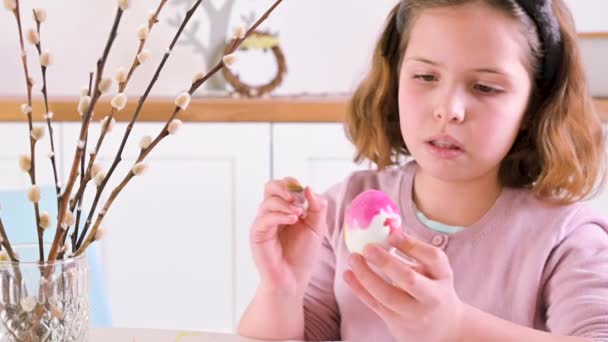 Egy kislány tojást fest húsvétra. Gyermek iskolás európai megjelenés és húsvéti tojás, dekoráció az asztalon. Világos belső tér és természetes felvételek. — Stock videók