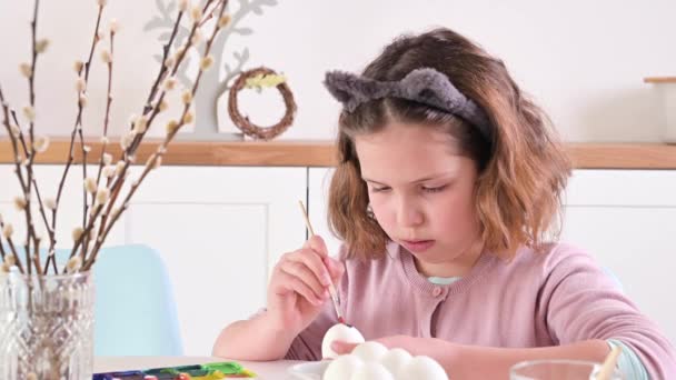 Uma menina pinta ovos para a Páscoa. Menino estudante de aparência europeia e ovos de Páscoa, decoração na mesa. Interior de luz e tiros naturais. — Vídeo de Stock