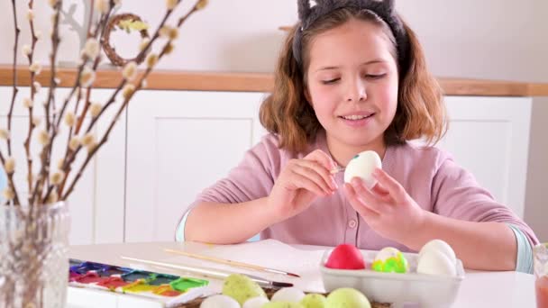 Une petite fille peint des œufs pour Pâques. Écolier enfant d'apparence européenne et oeufs de Pâques, décor sur la table. Intérieur léger et plans naturels. — Video