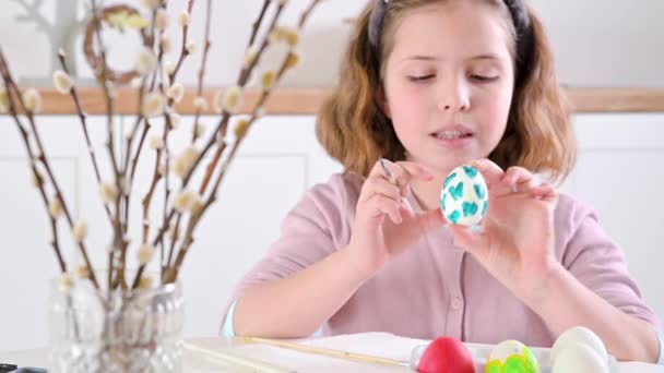 Een klein meisje schildert eieren voor Pasen. Kind schooljongen van Europees uiterlijk en paaseieren, decor op tafel. Lichte binnenkant en natuurlijke opnamen. — Stockvideo